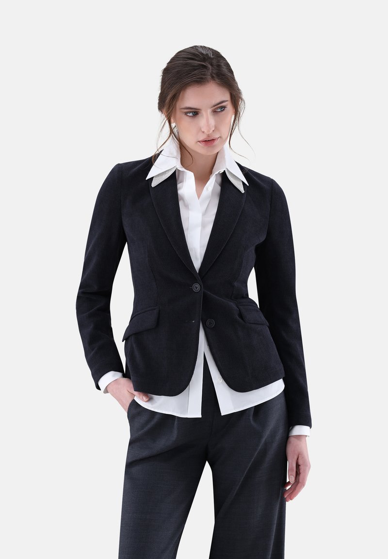 Schwarze Cordblazer mit strukturiertem Schnitt, zwei Knöpfen und Taschen, über einem weißen Button-up-Hemd mit kontrastierendem Kragen getragen.