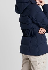 Tmavě modrá péřová bunda s kapucí, řaseným detailem v pase a bočními kapsami. Vyrobena z hladké, izolované látky.