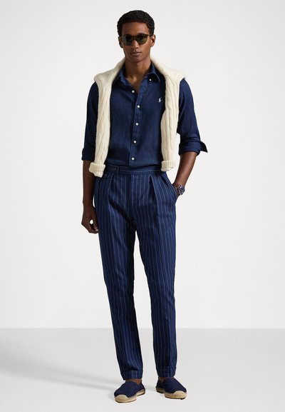 Melnā un baltā svītraina bikses ar krokām, apvienotas ar zilu lina kreklu un krēmkrāsas adītu vestu. Aksesuāri ietver tumšas saulesbrilles un espadrilles.