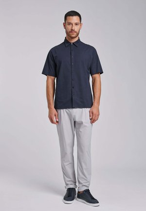 Mężczyzna stoi zwrócony frontalnie, ubrany w granatową koszulę na krótki rękaw z guzikami, jasnoszare spodnie i ciemne sportowe buty na jednolitym tle.