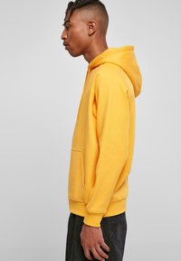 Sudadera amarilla de tela suave, con bolsillo frontal, capucha ajustable y puños acanalados, utilizada con pantalones oscuros.