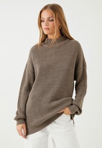 Sweter dzianinowy w ciepłym kolorze taupe, z wysokim dekoltem, luźnym krojem i prążkową strukturą. Długie rękawy z zawiniętymi mankietami.