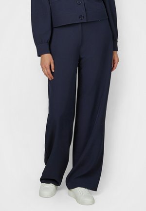 Person trägt marineblaue Hose mit weitem Bein, marineblauen Knopfjacke und weiße Turnschuhe, steht vor einem schlichten hellen Hintergrund.