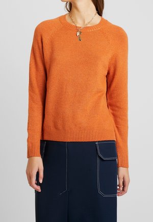 Personne portant un pull orange à manches longues et une jupe bleu foncé avec un détail de poche blanche cousue, accessoirisée d'un collier chaîne en or.