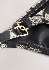 Černý horní díl bikin s květinovým vzorem v krémové barvě, vybavený zlatou sponou a hladkou texturou látky podél okrajů.