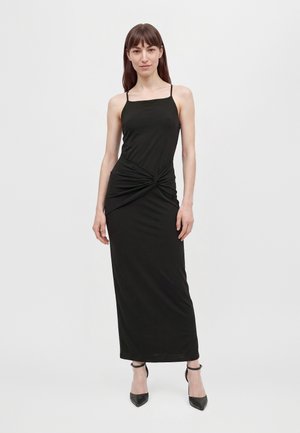 Le mannequin se tient devant un fond blanc uni, vêtu d’une longue robe noire à fines bretelles, froncée à la taille, et de chaussures noires à talons.