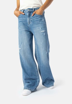 Helle blaue Schlagjeans mit abgenutzten Details, fünf Taschen und Knopfverschluss, getragen mit weißen Sneakers.