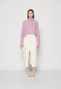 Suéter de cuello alto de color púrpura claro, falda crema de longitud media con una abertura frontal, y zapatos de tacón alto blancos. Cartera en una mano.