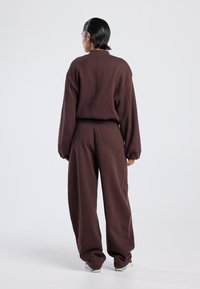 Persoon die met de rug naar de kijker staat, gekleed in een losse donkerbruine sweatshirt en bijpassende sweatpants, met wit-blauwe sneakers op een effen lichte achtergrond.