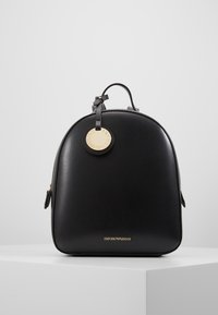 Mochila de cuero negro con forma redondeada, que presenta una etiqueta de logo en dorado, cierres de cremallera y un acabado texturizado. El nombre de la marca está grabado en la parte inferior.