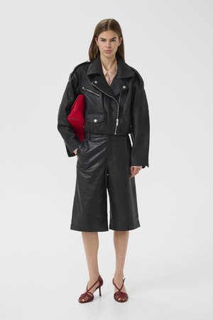 Kvinde iført sort læderjakke og culottes, holder en rød clutch, kombineret med lyserød bluse og burgunderfarvede hælede sandaler, stående på hvid baggrund.