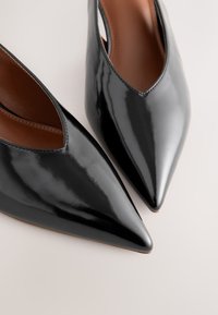 Mustad lakknahast kõrged kontsakingad teravate varvaste ja avatud külgedega, millel on sile ja läikiv viimistlus ning pruun sisemust vooder.