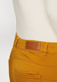 Gula bomullsbukser med en synlig läderlapp vid midjan. Funktioner inkluderar sömdetaljer och en bakficka.