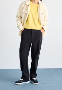 Krämfärgad fleecejacka, gul t-shirt, svarta byxor och svarta sneakers med vita sulor och krämfärgade snören. Tygernas texturer varierar.