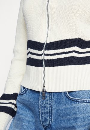 Pull en maille blanc avec des rayures noires et une fermeture éclair centrale, porté avec un jean bleu, montrant le torse et l'avant-bras.