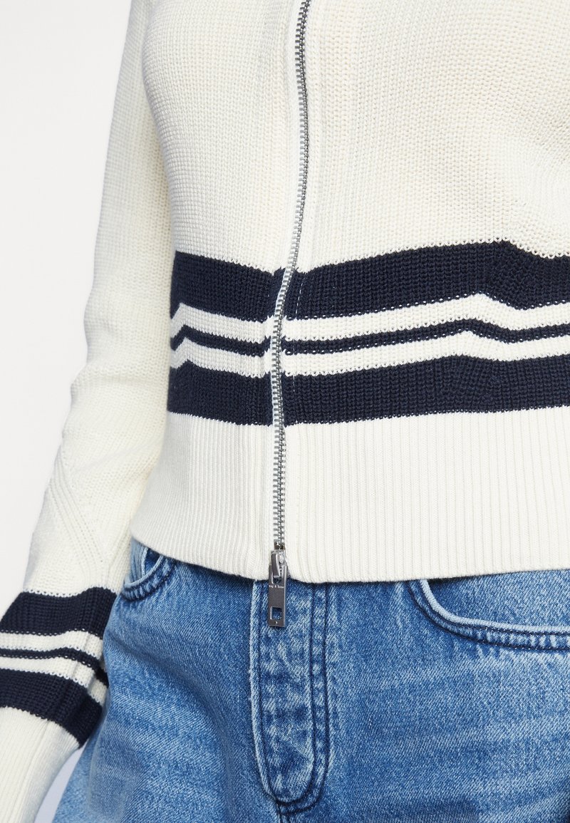 Pull en maille blanc avec des rayures noires et une fermeture éclair centrale, porté avec un jean bleu, montrant le torse et l'avant-bras.