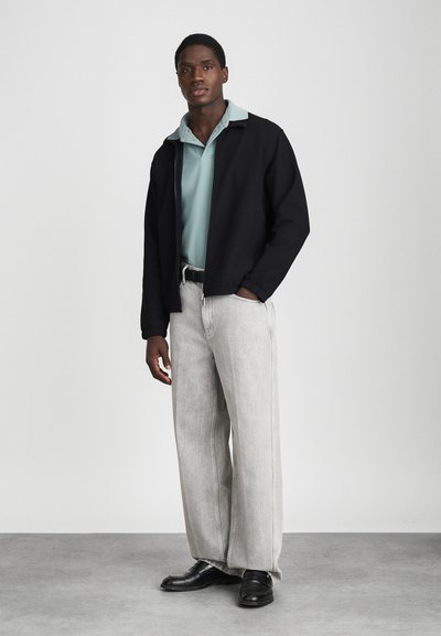 Čierna zipová bunda cez svetlomodrý polokošeľu, skombinovaná s svetlosivými širokými džínsami a čiernymi topánkami. Minimalistický dizajn, hladké textúry látok.