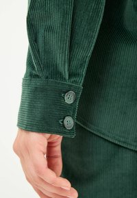 Groene corduroy overhemdmanchet met verticale ribbels, twee donkere knopen en een gestructureerde pasvorm. Geen extra elementen aanwezig.