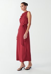 Červené midi šaty s jedním ramínkem, řasenými detaily a zavázaným pasem. Hladká tkanina doplňuje černé sandály na vysokém podpatku.