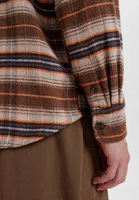 Bruine geruite flanellen overhemd met oranje, blauwe en witte strepen, met een knoopmanchet detail en een gestructureerde afwerking. Gelaagd over een bruine stof.