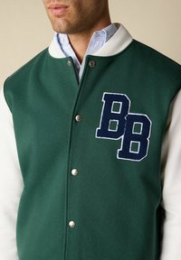 Man in een groene varsityjack met witte mouwen, zilveren knopen en een marineblauwe dubbele "BB" patch op de borst.