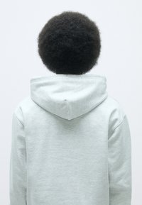 Felpa leggera grigia realizzata in morbido tessuto, dotata di cappuccio e spalle basse. La vista posteriore mostra una superficie liscia e un design minimalista.