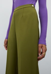 Olijfgroene wijde broek met hoge taille, voorzien van een zijrits, een gladde textuur en metalen accenten. Gepaard met een aansluitende paarse geribbelde top.