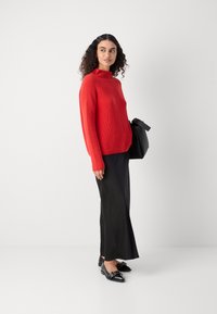 Roter gerippter Pullover mit hohem Kragen, kombiniert mit einem schwarzen engen Rock und schwarzen Schuhen mit Akzenten. Modell hält eine schwarze Tasche.