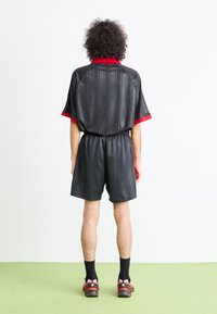 Una persona con capelli ricci sta in piedi di spalle, indossando pantaloni e maglietta atletici neri e rossi, con calzini neri e scarpe da ginnastica con dettagli rosa.