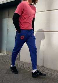 Rosa Kurzarmshirt, das über einem schwarzen Langarmshirt getragen wird; blaue Jogginghose mit einem orangefarbenen Emblem; schwarze Turnschuhe; läuft auf dem Bürgersteig.