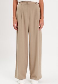 Pantaloni beige a gamba larga con vita alta, dotati di chiusura con bottone, pieghe e una texture morbida e liscia. Indossati con sneakers bianche.