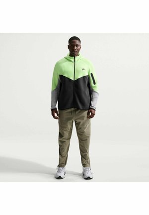 Uomo che indossa una giacca Nike con cerniera color verde neon, nero e grigio, pantaloni beige e sneaker bianche, in piedi davanti a uno sfondo bianco uniforme.
