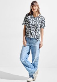 Grå og sort mønstret t-shirt med korte ærmer, parret med lyst blå brede jeans og hvide sneakers. Glat stofstruktur.