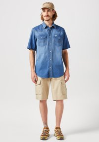 Camicetta in denim a maniche corte in blu chiaro e scuro, con due tasche sul petto e dettagli di cucitura, abbinata a pantaloni cargo beige e scarpe da trekking.