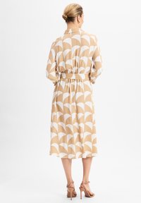 Beige jurk met witte gebogen patronen, lange mouwen, elastische taille en knielengte. Gecombineerd met beige sandaaltjes.