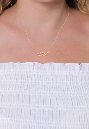 Collier en or avec un pendentif en forme de soleil porté autour du cou d'une femme, visible au-dessus d'un haut bustier blanc texturé.
