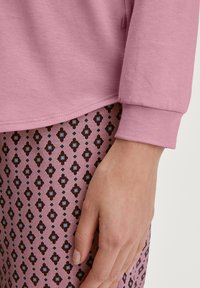 Lichtroze lange mouwtop met een afgeronde zoom die overlapt met een paar patroonbroeken met geometrische vormen in roze, bruin en blauw.