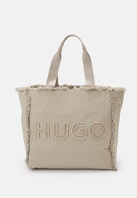 Beige Tragetasche mit ausgefransten Kanten, die ein gesticktes "HUGO"-Logo in einer hellen Farbnuance zeigt. Robustes Material und doppelte Stoffgriffe.