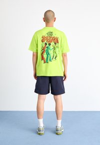 Neongrön t-shirt med en stor grafisk illustration på baksidan, som föreställer tre figurer som dansar med texten "de nya originalen."
