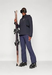 Isik kannab musta suusajakki, siniseid suusapükse, musti suusahindeid ning suuri musti suusaglaase, hoides püsti suuskade paari.