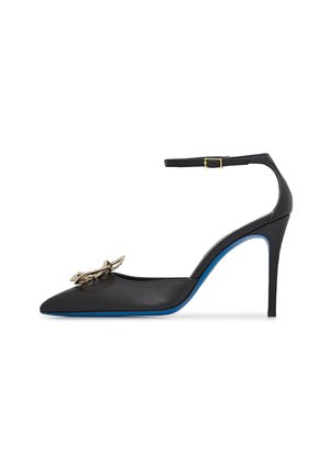 Scarpa con tacco alto in pelle nera, con punta affilata, cinturino alla caviglia e dettaglio a fiocco con accenti dorati. Presenta una suola blu e un design elegante.