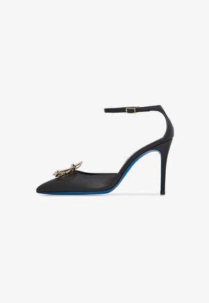 Scarpa con tacco alto in pelle nera, con punta affilata, cinturino alla caviglia e dettaglio a fiocco con accenti dorati. Presenta una suola blu e un design elegante.