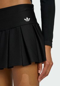 Čierna skladaná sukňa z texturovanej látky, s vysokým pásom a malým bielym logom Adidas vpredu. Obsahuje aj detail so zipsom.