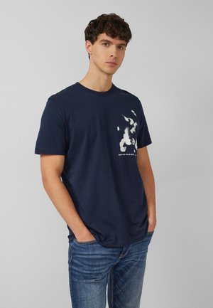 Młody mężczyzna z krótkimi kręconymi ciemnymi włosami, ubrany w granatowy T-shirt z grafiką i niebieskie dżinsy, stojący z rękami w kieszeniach na szarym tle.