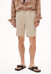 Shorts beiges à coupe droite, avec des passants de ceinture et une fermeture à bouton à l'avant. Portés avec une chemise à manches longues à carreaux marron et des sandales noires.