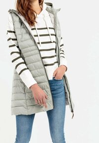 Lichtgrijs, mouwloos pufferbodywarmer met zijzakken, doorgestikte textuur en ritssluiting, gedragen over een witte hoodie met groene strepen.