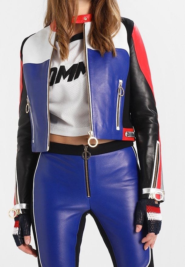 Femme portant une veste en cuir bleu, rouge, noir et blanc avec un pantalon assorti, un haut en maille blanc court, et des gants sans doigts.