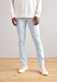 Világoskék, slim-fit farmernadrág, bézs pulóverrel és fehér sportcipőkkel párosítva. A farmernadrág sima felületű és minimalista dizájnnal rendelkezik.