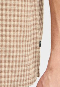 Brown and beige checkered fabric with a textured surface, featuring a side tag, displaying a casual shirt design and soft cotton material.