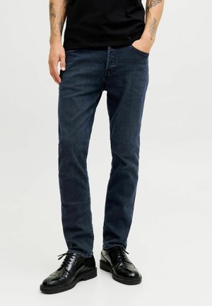 Homme portant un jean slim bleu foncé, des chaussures habillées noires et une chemise noire, debout avec une main dans la poche.